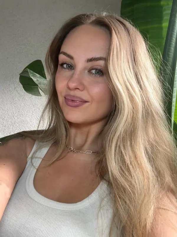 A woman with long blonde hair smiling softly beside a houseplant.