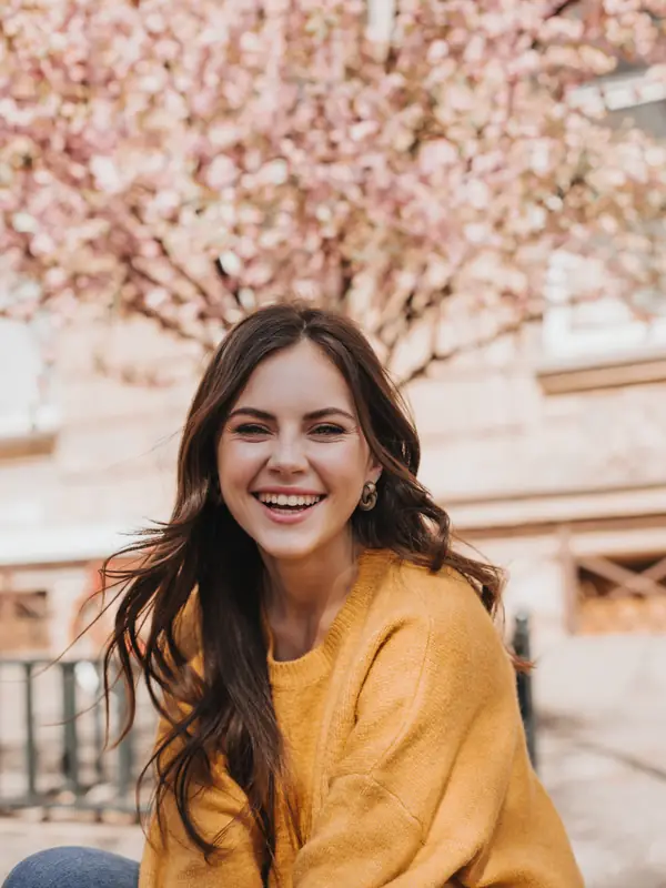 A smiling woman in a mustard jumper under a blossoming cherry tree.