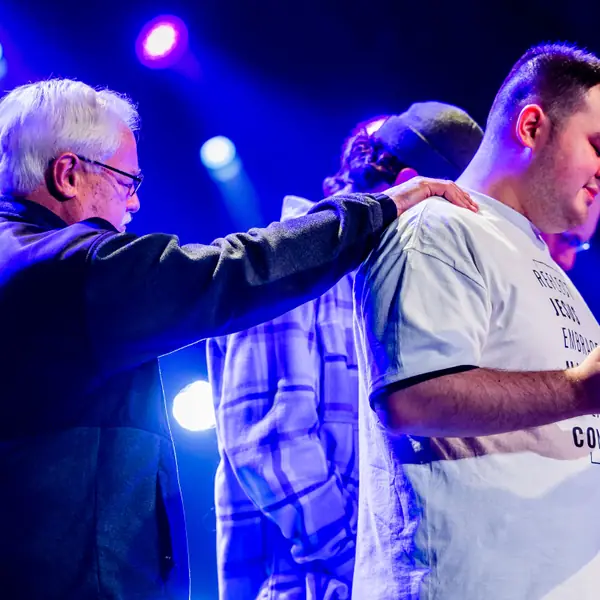 Two men praying together with a hand on the shoulder, under stage light.